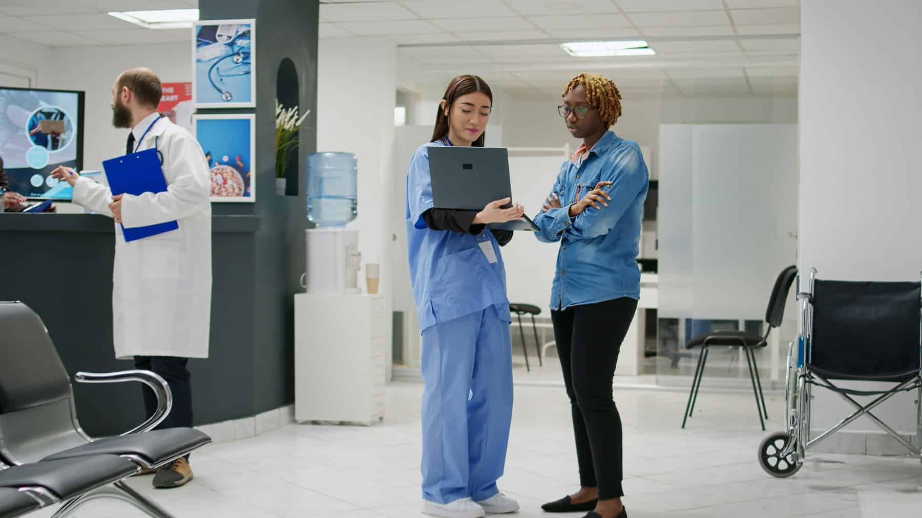 Healthcare professional in blue scrubs reviewing a laptop with another person in a medical clinic waiting area.