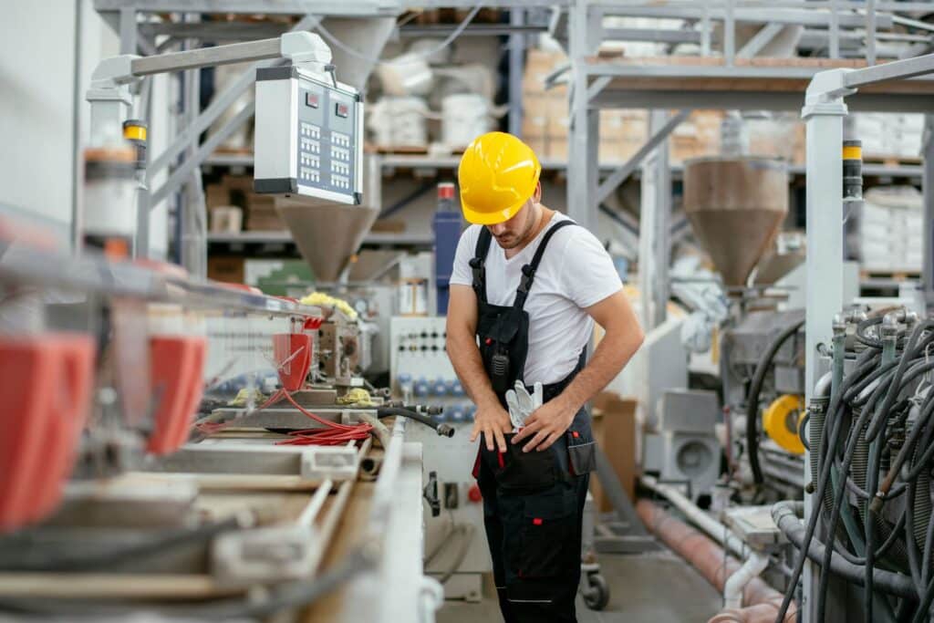 Factory worker wearing a yellow hard hat and black overalls beside industrial machinery and control panels.