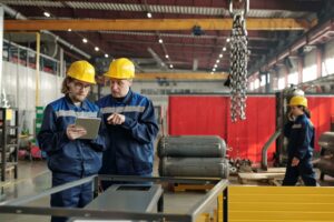 Two manufacturing workers in yellow hard hats reviewing a tablet beside equipment on a factory floor.