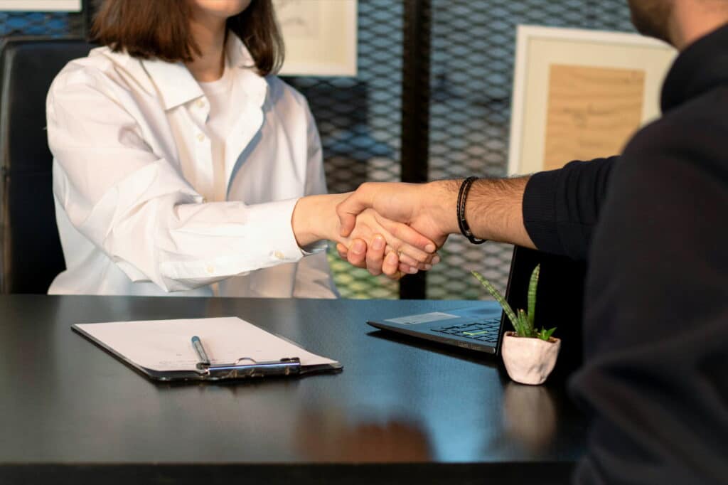 Two people shake hands across an office desk during a professional meeting, with a laptop, clipboard, and small potted plant nearby.