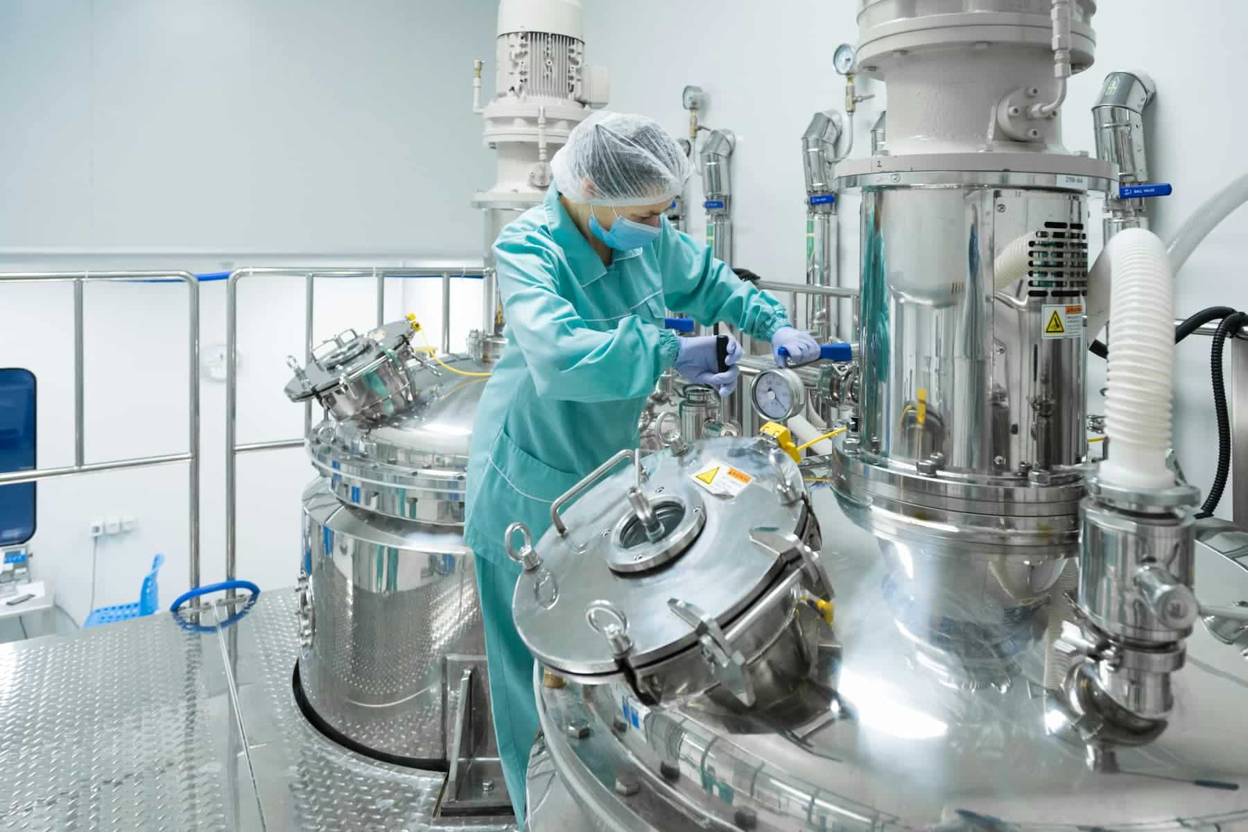 Technician wearing a mask, hairnet, and gloves adjusts stainless steel processing equipment in a clean manufacturing facility.