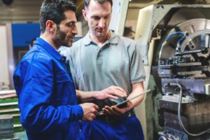 Two manufacturing workers review a tablet beside an industrial metalworking lathe.